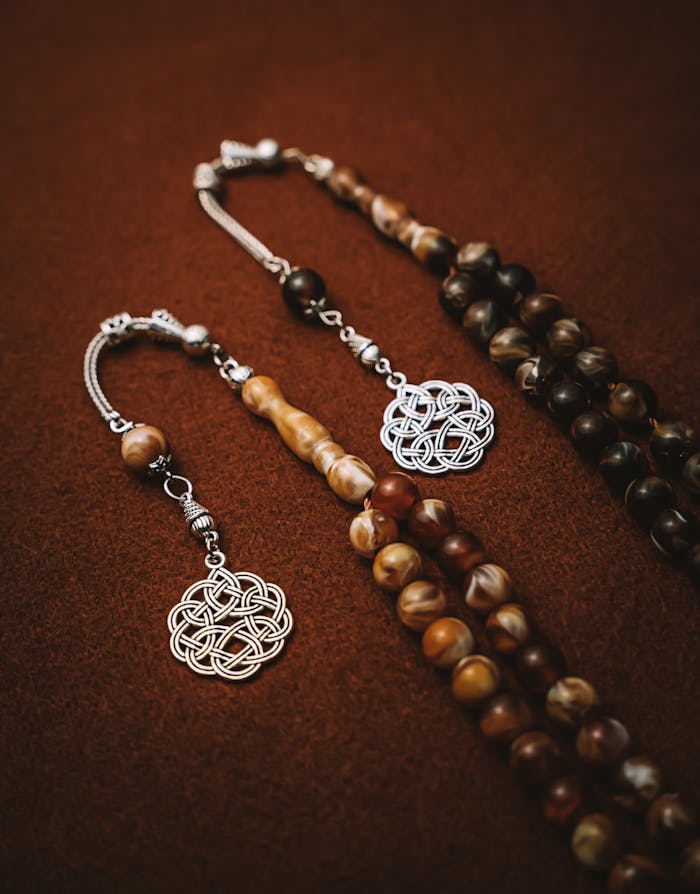 Home Elegant brown prayer beads with intricate silver pendants displayed on a textured brown surface.